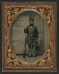 [Unidentified soldier in Union uniform and forage cap with bayoneted musket in front of painted backdrop showing military camp and American flag] (LOC)