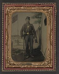 [Unidentified soldier in Union uniform with bayoneted musket, canteen, cartridge and cap boxes in front of painted backdrop showing military camp with American flag] (LOC)