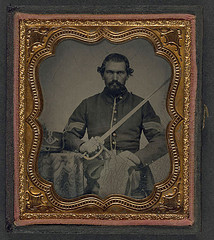 [Unidentified soldier in Union infantry sergeant's uniform holding a model 1840 non-comissioned officer's sword next to table with Company H, 6th Regiment Hardee hat on top] (LOC)