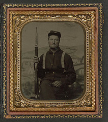 [Unidentified soldier in Union uniform with carbine in front of painted backdrop showing military camp scene] (LOC)