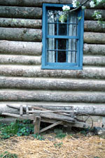 View of the side of a log structure, with a wooden wheelbarrow below a blue window.