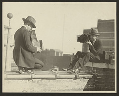 [Two photographers taking each others' picture with hand-held cameras while perched on a roof] (LOC)