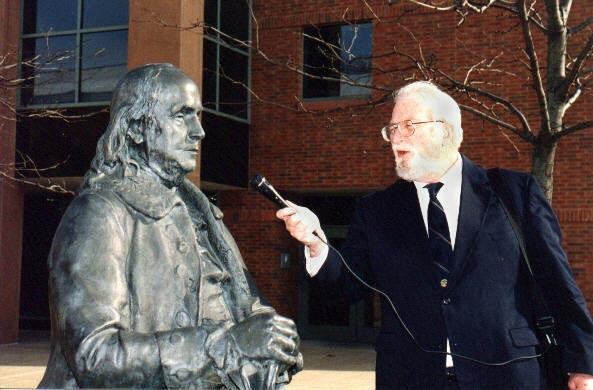 A man holding a microphone pretends to interview a statue of Benjamin Franklin