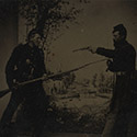 Two soldiers in Union uniforms posing with weapons in front of painted backdrop showing landscape with camp