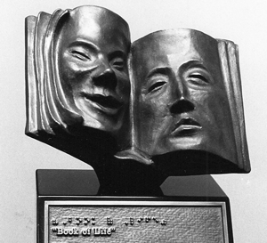 Bronze sculpture of an open book with the impressions of a happy face on a left-hand page and a sad face on the right-hand page. Beneath the book sculpture is a plaque that reads both in braille and text: “Book of Life”