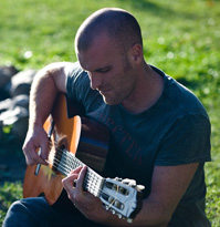 Young adult male playing his acoustic guitar outdoors on a lawn.