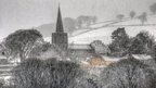 A village scene, with a church, trees, fields and houses is white in the snow. Snow is falling in this shot, making it look slightly blurry.
