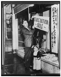 A Japanese-American man hanging a sale sign on a shop window. A young boys stands near him.