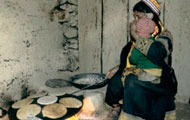 Woman squats, toddler on her lap, cooks large pan full of flat bread over indoor cookstove, flames visible beneath pan