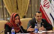 Woman wearing head scarf and man seated at conference table, woman speaks into microphone, Iranian flag in background