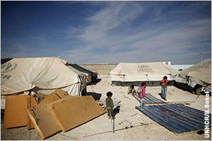 Bright blue sky over desert terrain, several tents in foreground with children playing amongst them (UNHCR/B.Sokol)