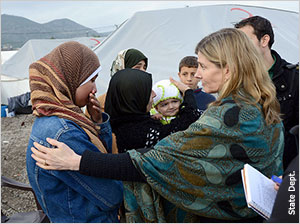 ليندبورغ (صورة جانبية) تضع يدها على كتف سيدة سورية وهي تتحدث معها وتظهر وراءهما صفوف من الخيام وتلال (مصور الخارجية الأميركية)