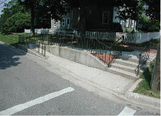 Photo shows a sidewalk in a residential neighborhood with stairs and a ramp down to a much-lower street level.