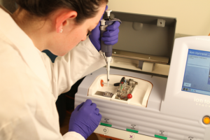 Device on benchtop in lab, featuring open lid for placement of microarray chip and to the right, a read-out screen. At bottom of device are test tubes containing liquid samples. 