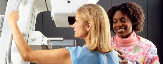 An African-American female technician positions a Caucasian woman at an imaging machine to receive a mammogram.