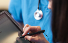 A woman uses a stylus on a tablet with person wearing a stethoscope visible in the background