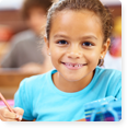 A young girl in a blue tee shirt smiling at the camera as she holds a pencil in her hand. Other students appear to be in the background.