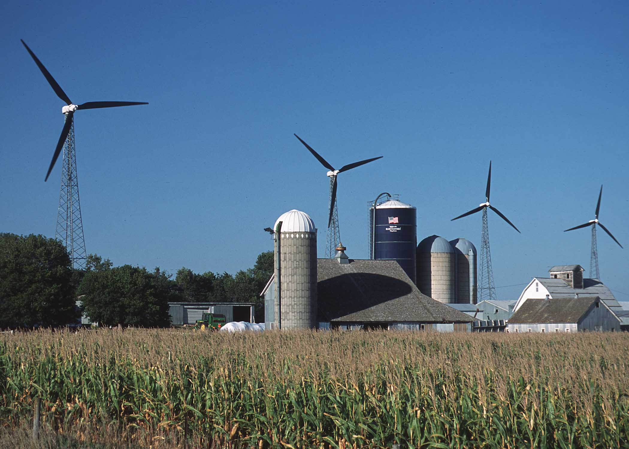 Picture of silo, green fields, with several modern wind generators in background, blue sky.