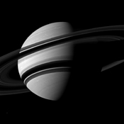 The Cassini spacecraft takes an angled view toward Saturn, showing the southern reaches of the planet with the rings on a dramatic diagonal. The moon Enceladus appears as a small, bright speck in the lower left of the image.