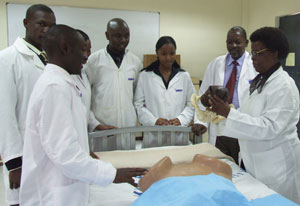 Group of medical students in white lab coats stands around hospital bed, woman leading discussion points to model she holds