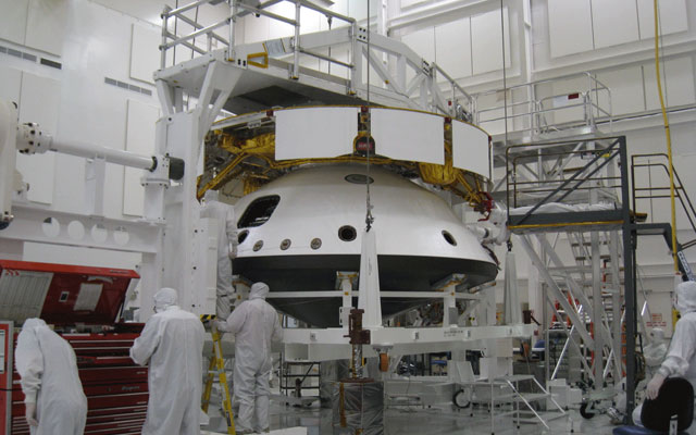 This image shows the cruise structure (the 'ring' on top) and one part of the aeroshell (the backshell with the rover inside) flipped over and the bottom of the aeroshell (the black heat shield) being added. There are four mission team members in the foreground.