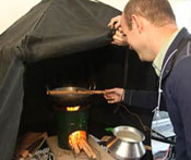 Man holds instrument to measure emissions over lit wood-burning stove, which burns under a hood
