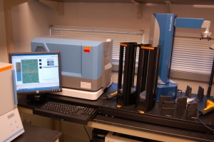 From left to right, flat screen monitor showing bead scan with green and red dots of genome; light blue sequencing unit; tray holder for microarray plates containing genome samples