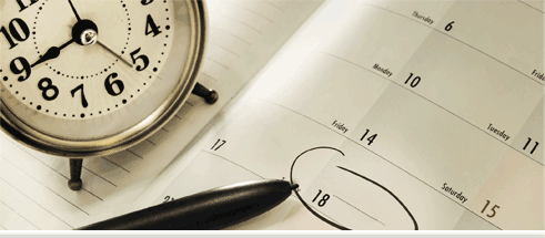 A round, manual clock and a pen on top of a calendar.