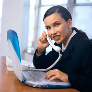 Photograph of a young woman doing research on her laptop computer while talking on the phone.