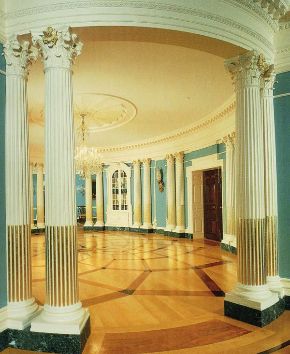 The Treaty Room at the State Department with curving blue walls, inlaid wooden floor, white columns, book cabinets, and chandelier. 