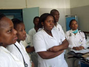 Group of 7 African health care workers in white lab coats in exam room listen attentively to speaker, not pictured