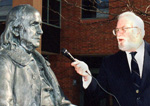 A man holding a microphone pretends to interview a statue of Benjamin Franklin