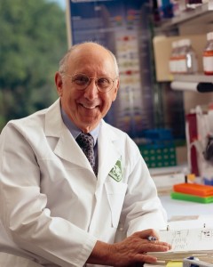 Image of caucasian man, wearing glasses, a light purple shirt, dark purple tie, and a white lab coat, sittling in a lab