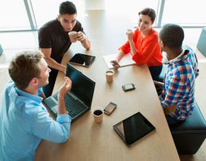 Photograph of diverse young adults talking over coffee and laptop computers.