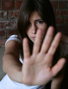 Photograph of  a teen girl standing with her back against a brick wall while holding up her hand in self-defense,
