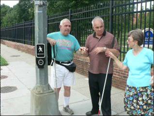 Two men stand beside a pole on a street corner. One has his arm on top of an APS pushbutton and his other hand under the hand of the second man. A woman is to their right gesturing. APS are visible on two sides of the pole.