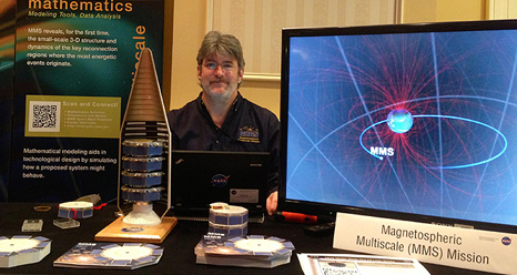 Craig Tooley sits next to a model of the spacecraft tucked into their launch rocket and a television showing the orbit of MMS as it travels through Earth's magnetic fields.