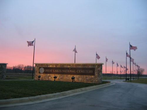 An image of the cemetery's front gate at dusk. Two rows of American Flags line both sides of the road as it leads onto the cemetery grounds.