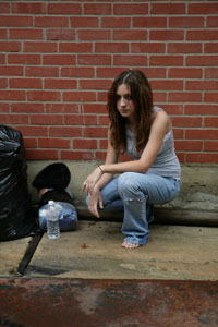 Photograph of a teen girl crouching on the sidewalk, wearing torn jeans and no shoes.