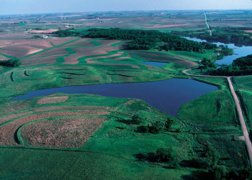 Low angle aerial photograph showing a blue lake behind a dam and green fields.
