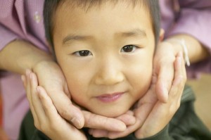 The hands of an unseen person tenderly frame the face of a young Asian boy.