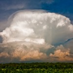 Huge white and orange-tinted clouds against blue sky above green landscape.