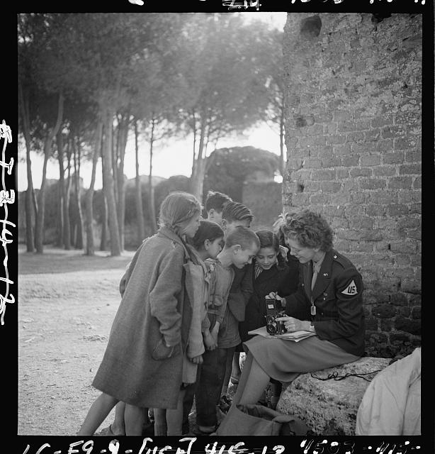 [Toni Frissell, sitting, holding camera on her lap, with several children standing around her, somewhere in Europe]