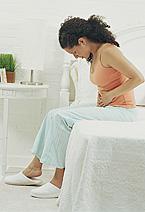 Woman sitting on bed holding stomach, head bowed.