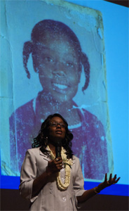 Photograph of Tonier Cain speaking with a photograph of her as a child in the background.
