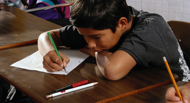 Child with head down writing on a piece of paper in classroom.