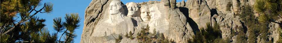 Mount Rushmore, Washington, Jefferson, T. Roosevelt, Lincoln framed by ponderosa pine trees under a bright blue sky.