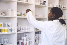 Photo of a woman in a white smock taking bottles of drugs off a shelf in a pharmacy.