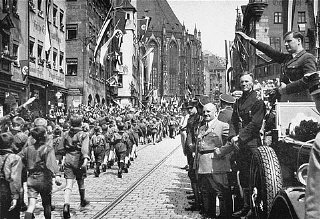 ہٹلر یوتھ کے ارکان اپنے لیڈر کے سامنے مارچ کرتے ہوئے، بالڈور وان شیراخ (دائيں طرف، سلامی دے رہا ہے) اور جولیئس اسٹرائخر سمیت دوسرے نازی اہلکار۔