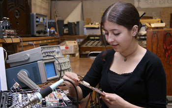 Image of a young woman holding a circuit board and next to table with electronics.